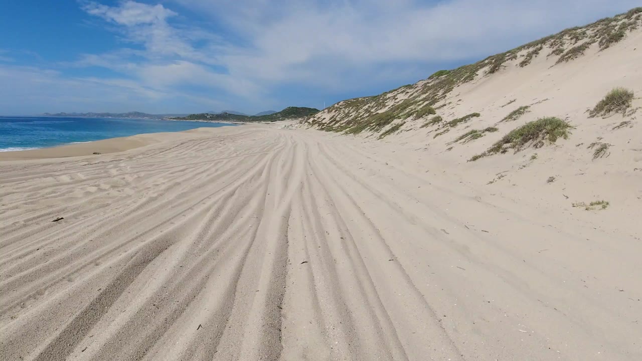 Opening view of a Los Cabos beach with ridged sand tracks leading forward, blue water on the left, scrub-covered dunes on the right, and no visible people.