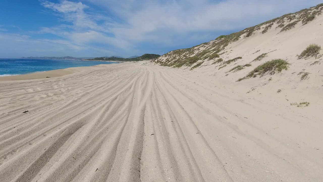 Second view along the Los Cabos shore, with deeper tire-like ridges in the pale sand, calm blue sea to the left, dunes to the right, and no visible people.