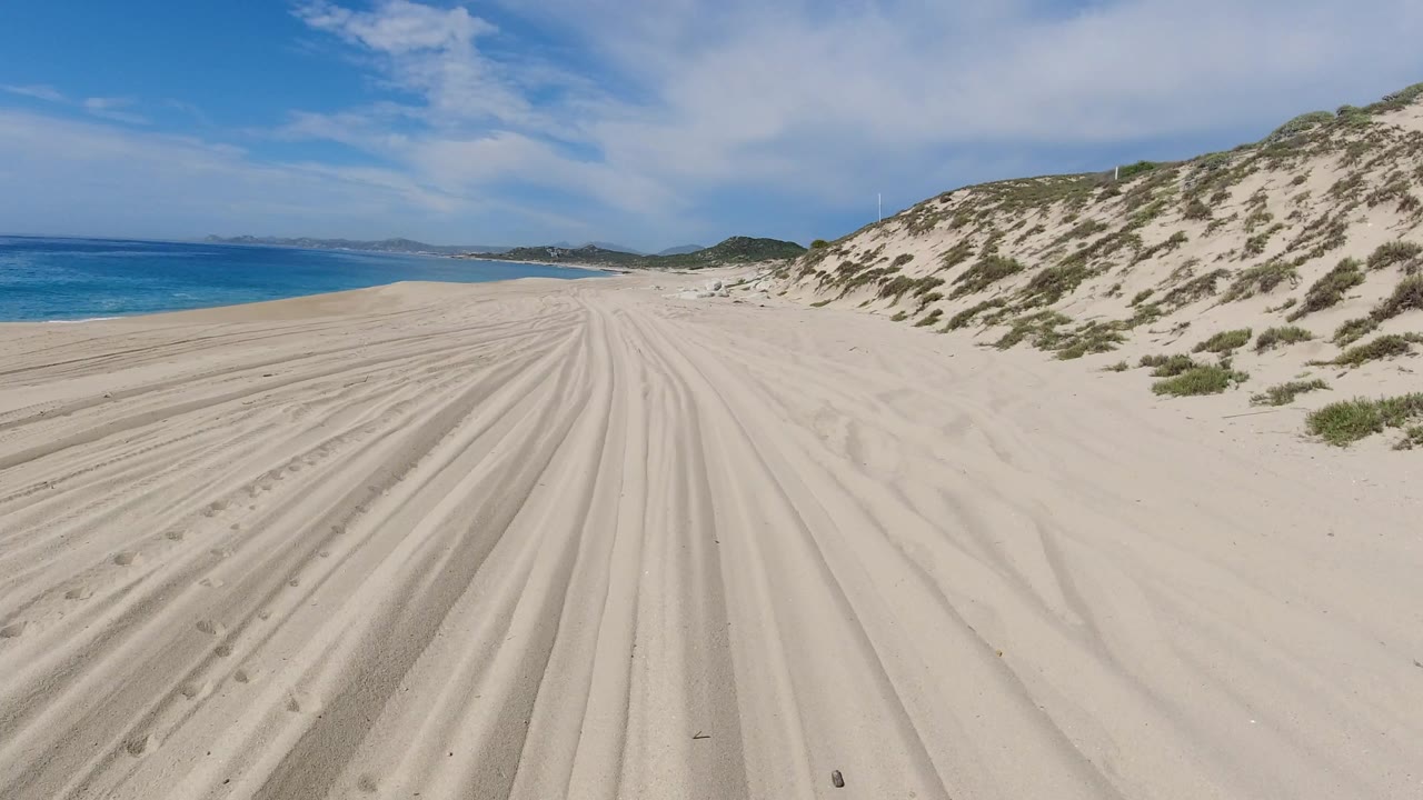 Los Cabos shoreline view with parallel marks in the sand centered in the foreground, calm blue water to the left, dune slopes to the right, distant landforms, and no visible people.