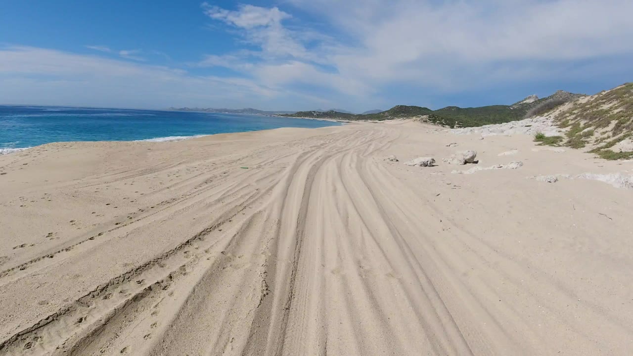 Wide Los Cabos beach view at the start of the clip, with tire tracks crossing pale sand toward distant hills, blue ocean on the left, dunes on the right, and no visible people.