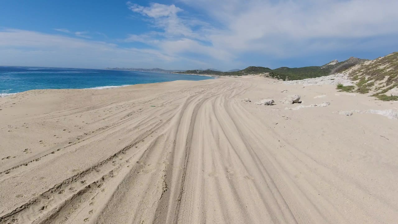 Forward-facing Los Cabos beach frame with parallel tire ruts in the sand, turquoise water along the left edge, pale dunes and rocks to the right, and no visible people.