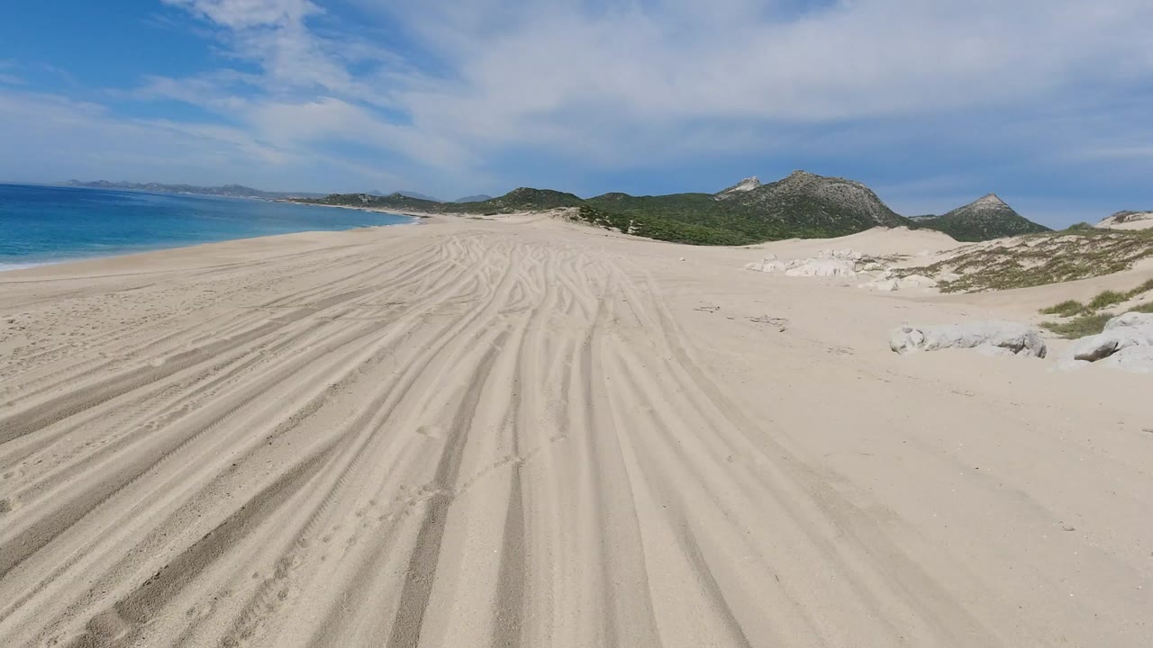 Los Cabos, Mexico sand tracks lead toward blue ocean on the left and rocky green hills under broken cloud, with no visible people.