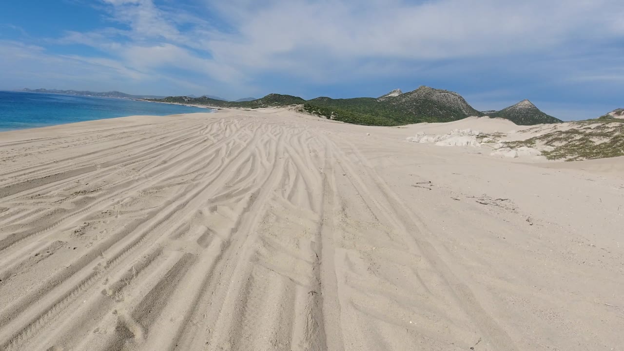Open beach dunes with curved tracks, blue water, and mountain backdrop.