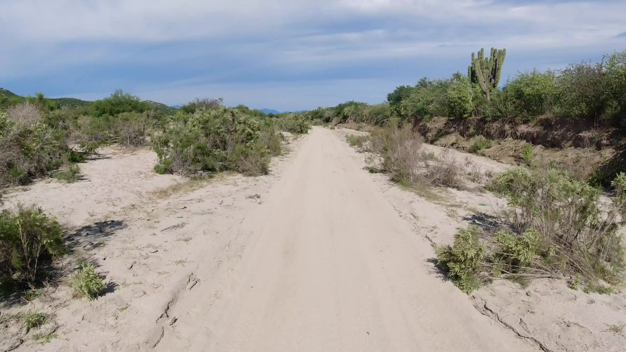 The Los Cabos dirt path stays centered as low green brush fills both sides and distant hills sit beneath a partly clouded sky, with no visible people.