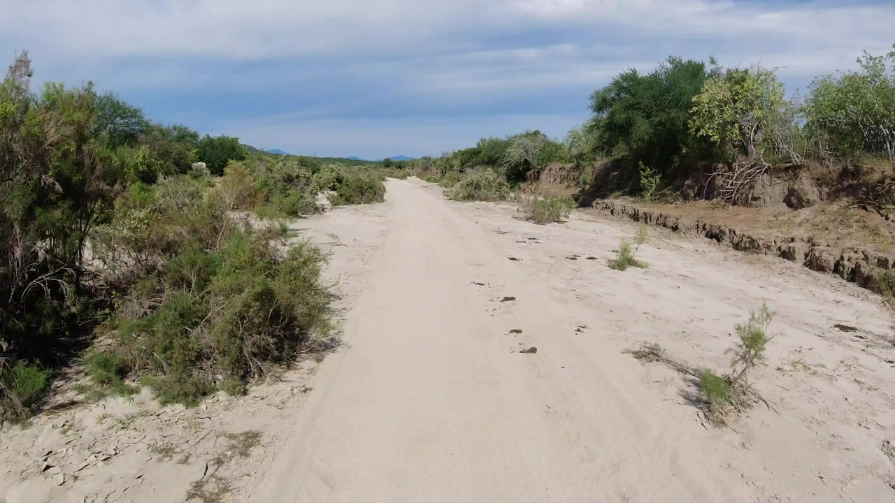Opening frame of a pale sandy road in Los Cabos, Mexico, bordered by green scrub and low eroded banks under a blue sky, with no visible people.