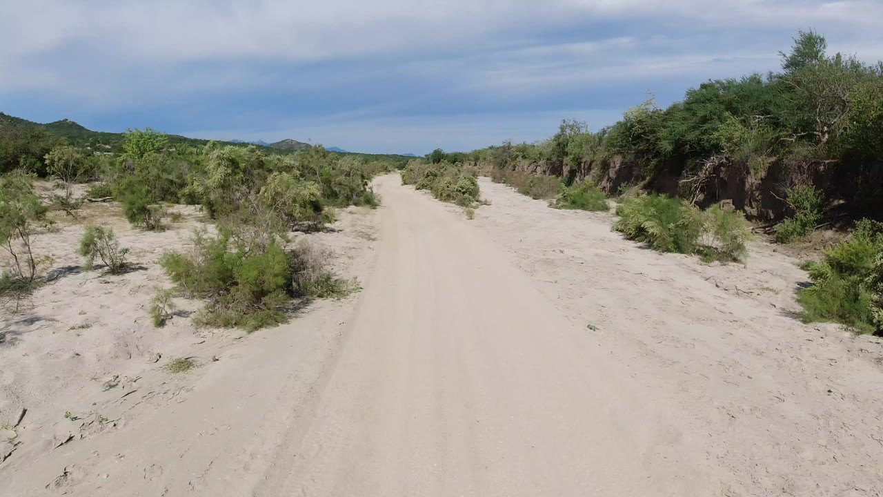 Opening view of a sandy dirt road in Los Cabos, Mexico, bordered by green shrubs and low hills in the distance, with no visible people.