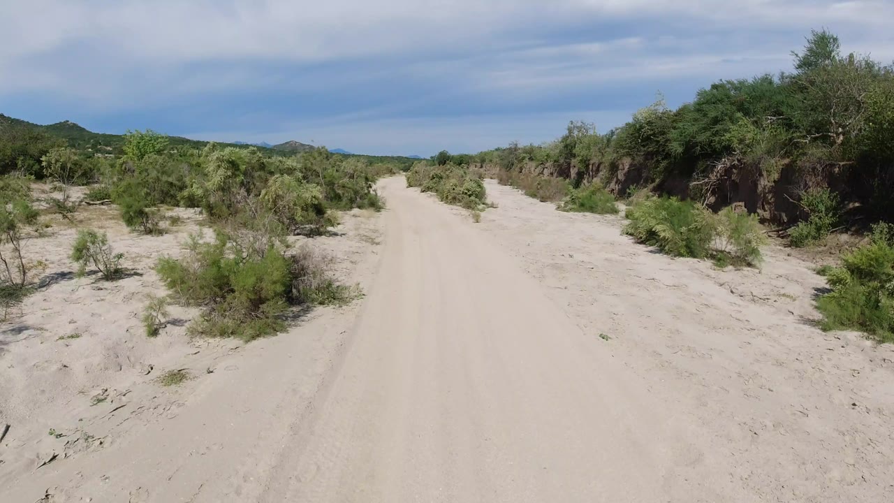 A later view down the pale dirt road in Los Cabos, Mexico, with scrub vegetation close to both sides and distant hills beneath thin clouds, with no visible people.