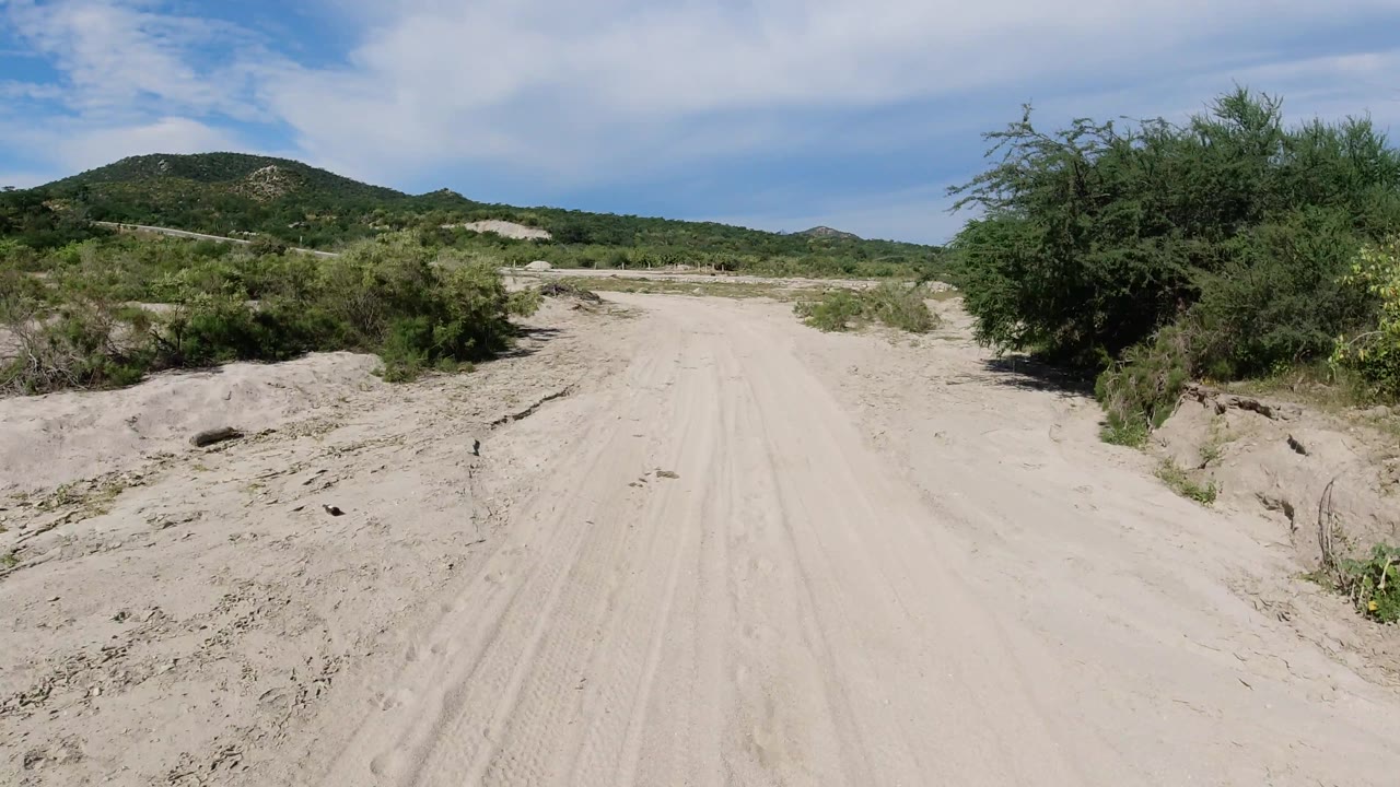 Sandy track curving past brush with hills and cloudy blue sky.