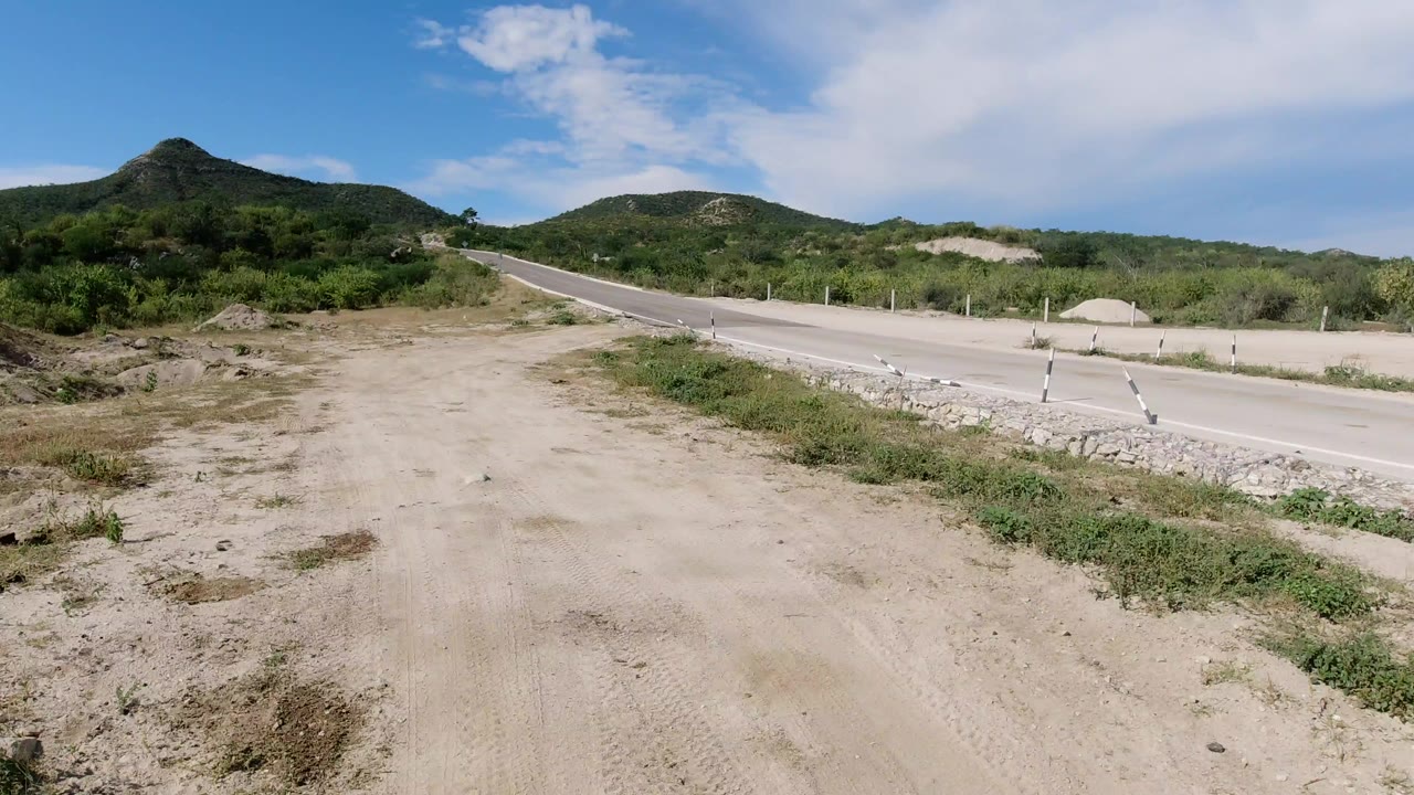 Wide elevated view in Los Cabos, Mexico, showing a sandy dirt track beside a paved road, green hills in the background, blue sky overhead, and no visible people.