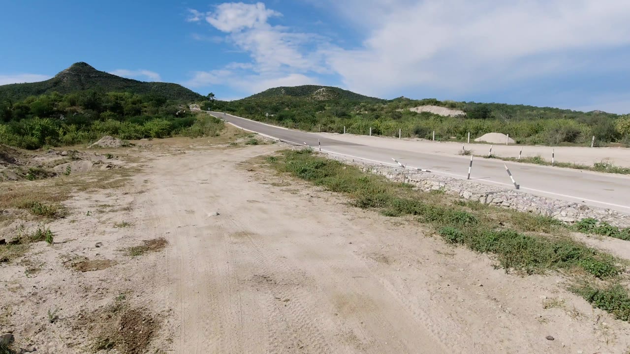 Elevated Los Cabos roadside view with pale dirt in the foreground, a paved road crossing the midground, brush-covered hills beyond it, and no visible people.