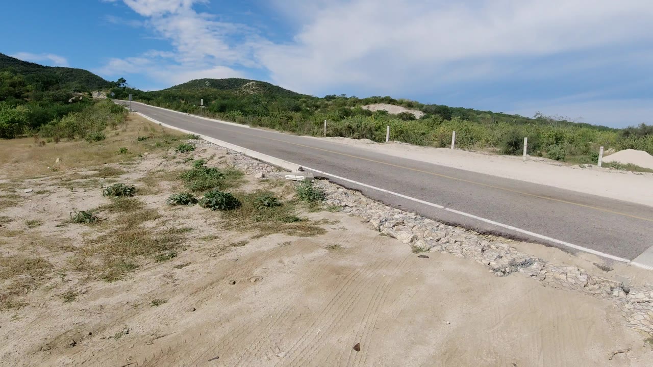 An empty paved road in Los Cabos runs diagonally past sandy shoulder, scrub vegetation, low green hills, and a blue sky with white clouds, with no visible people.