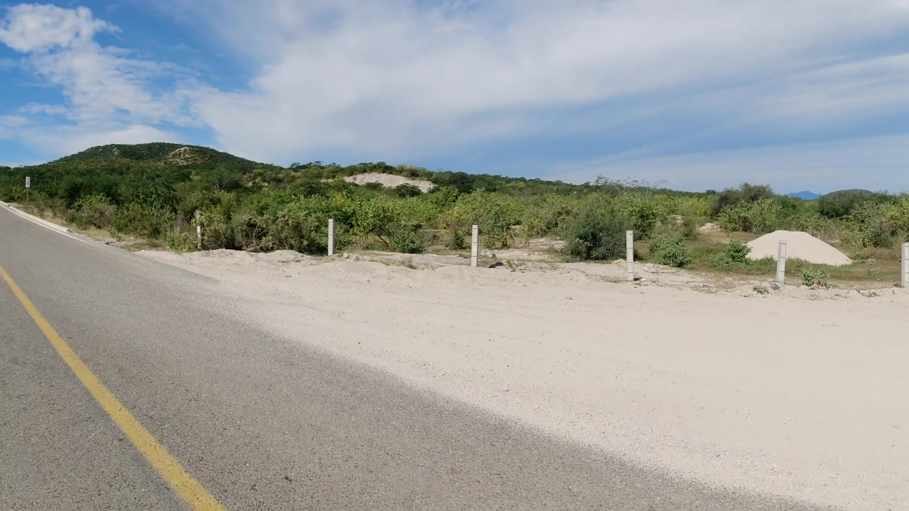 Paved road edge beside sandy scrub and low green hills