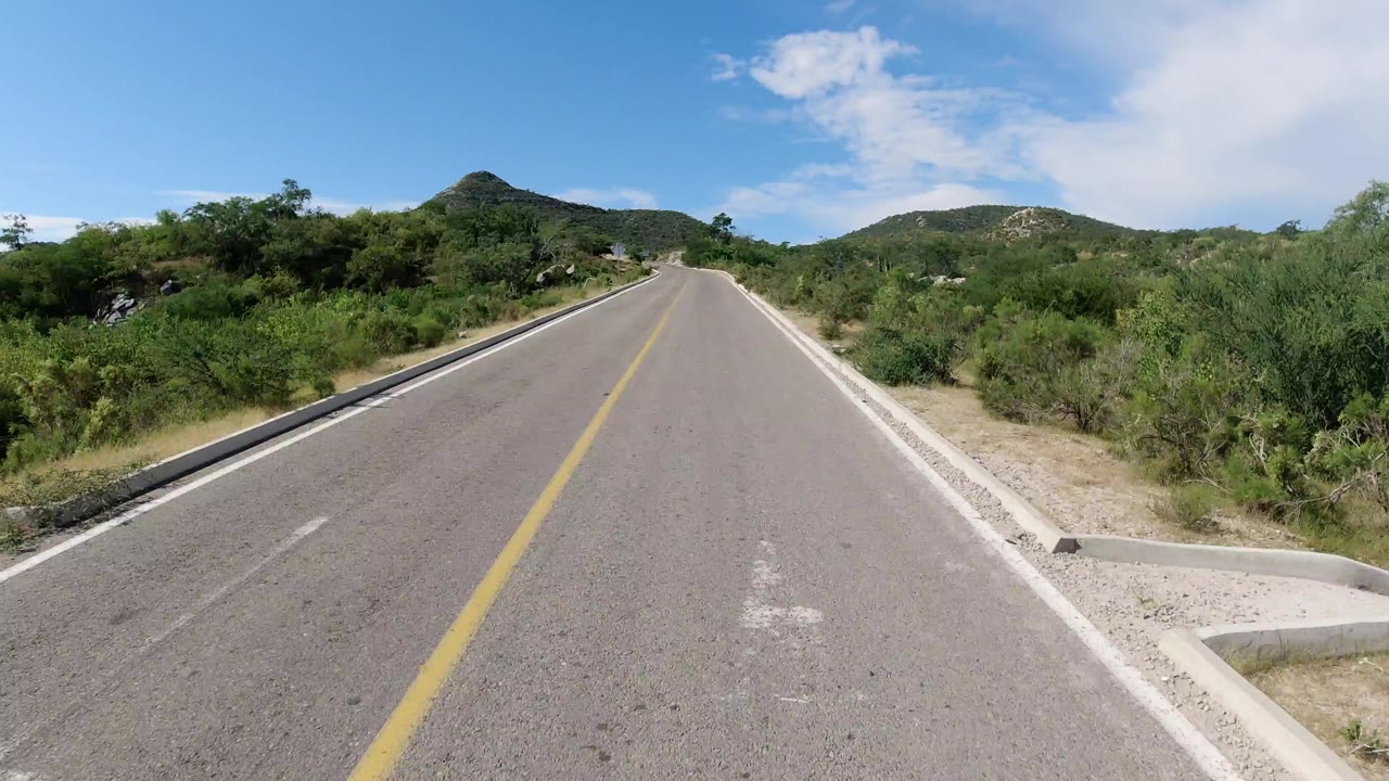 Forward view down an empty Los Cabos road with a yellow center line, green roadside bushes, low hills, blue sky, scattered clouds, and no visible people.