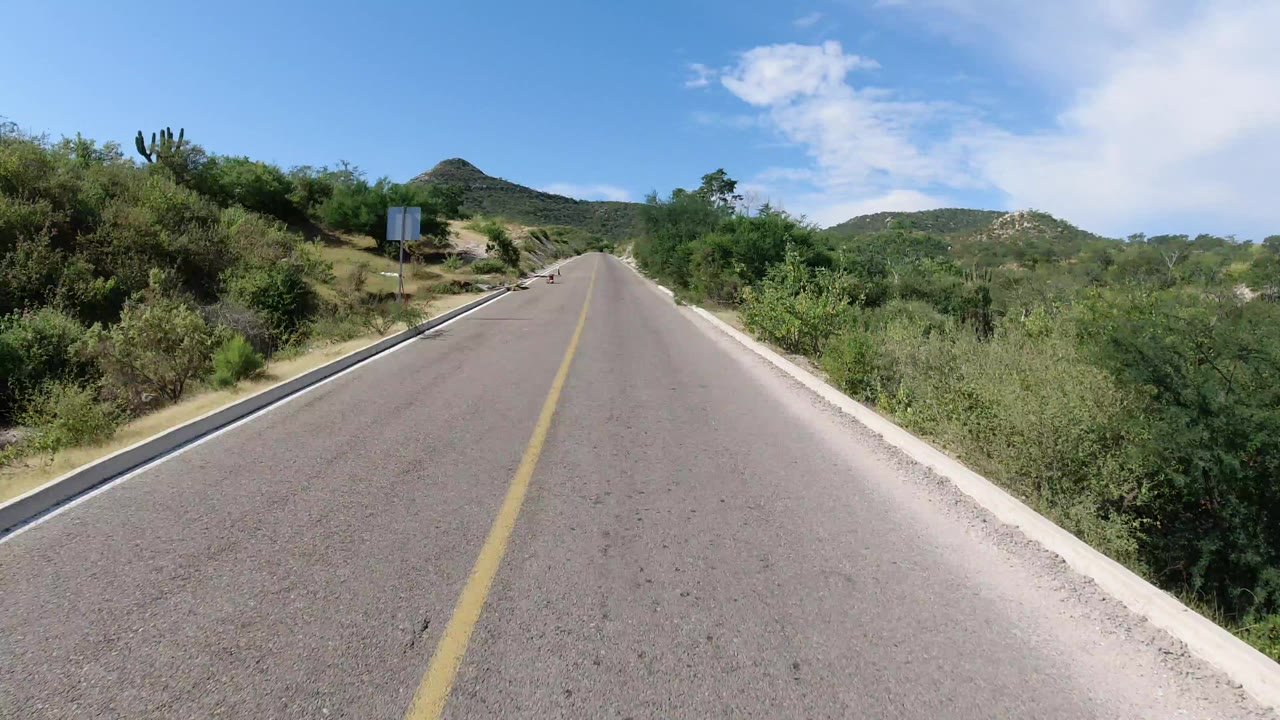 Empty paved road leading toward low hills under blue sky.