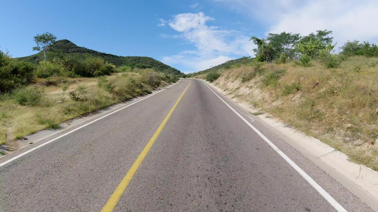 The road continues through Los Cabos, Mexico with a sandy raised shoulder at right, dense shrubs at left, green hills ahead, and no visible people.