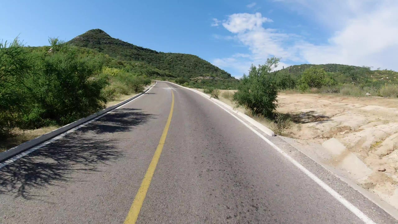 Los Cabos, Mexico road view with a yellow center line, roadside shrubs, green hills ahead, bright sky, and no visible people.