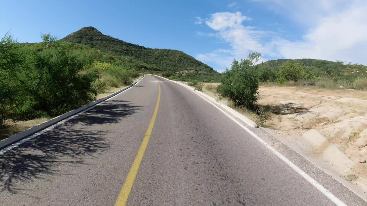 A low road-centered view in Los Cabos, Mexico shows the lane line pulling toward a rounded green hill, with dry shoulder and no visible people.