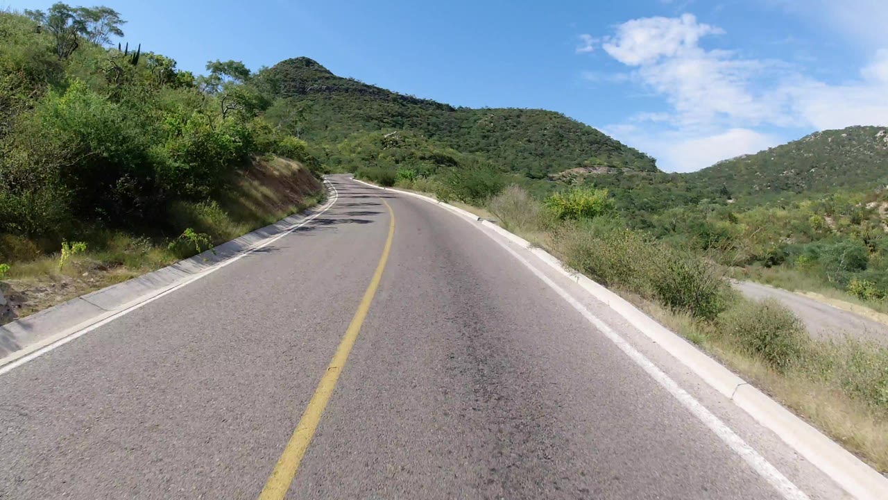 A Los Cabos mountain road continues uphill between roadside vegetation, with a cut bank on the left, green hills in the distance, blue sky, and no visible people.