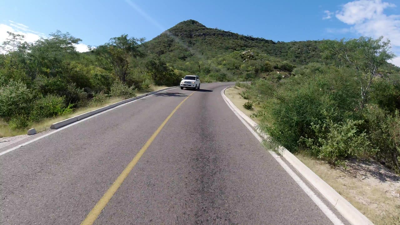 Curving road with a white vehicle ahead and green hills beyond.