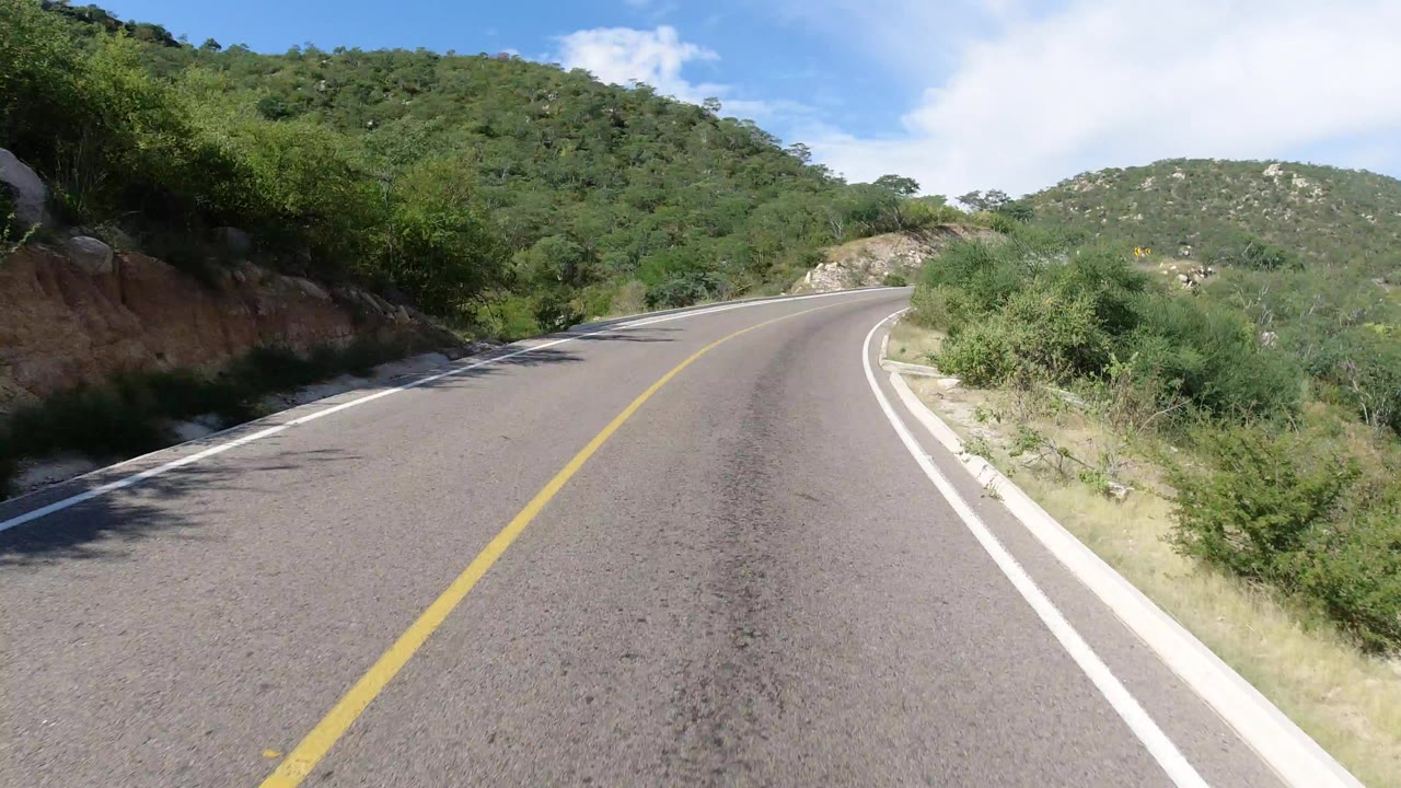 A Los Cabos, Mexico road curves ahead between roadside brush and green hills under bright blue sky, with no visible people.