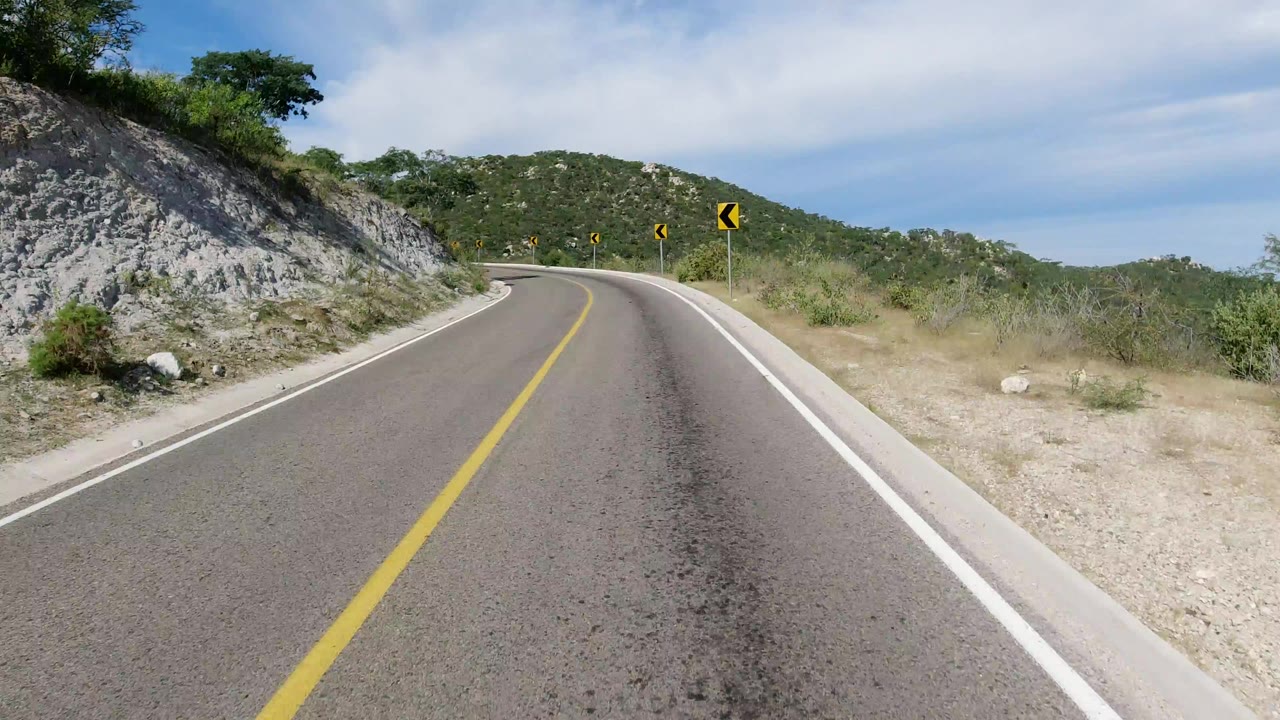 Opening view of an empty paved road in Los Cabos, Mexico, bending left past rocky hillside, scrub vegetation, yellow lane lines, and distant chevron signs, with no visible people.
