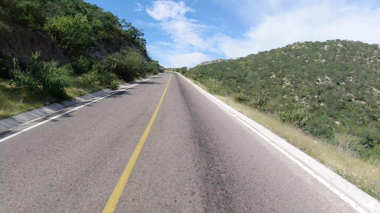 Forward road view in Los Cabos, Mexico, with an empty two-lane highway, scrub-covered hills, rocky left shoulder, and no visible people.