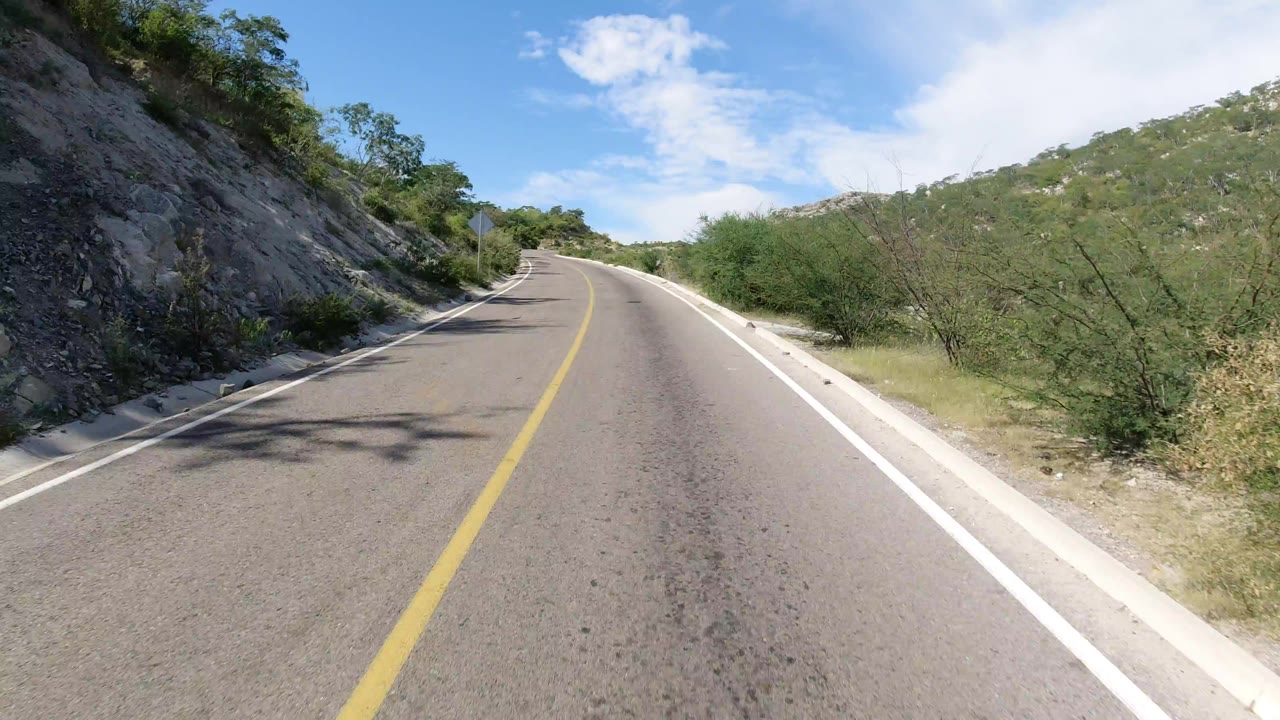 Curving paved road beside rocky hillside and green brush.