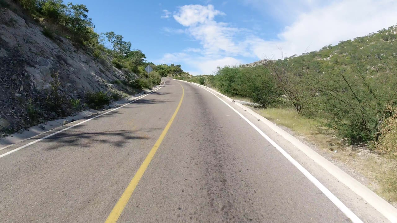 Empty Los Cabos road seen from the center of the lane, with asphalt texture in front, a rocky bank on the left, scrub on the right, distant hills, and no visible people.