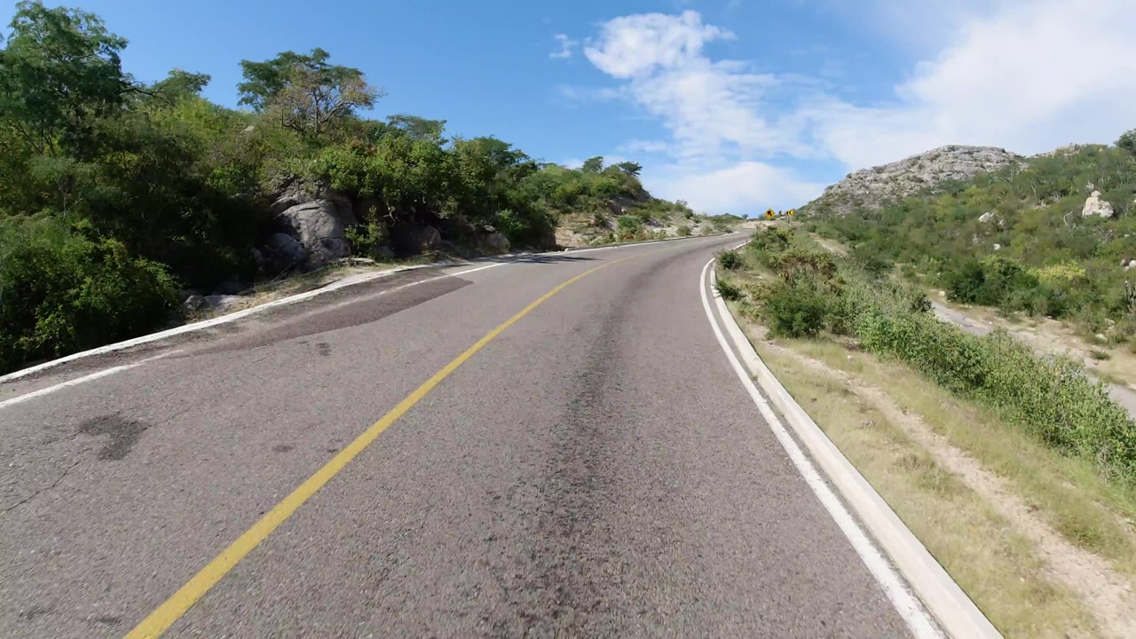 The Los Cabos roadway continues toward a left bend, framed by green brush, pale rocks, and open sky, with no visible people.