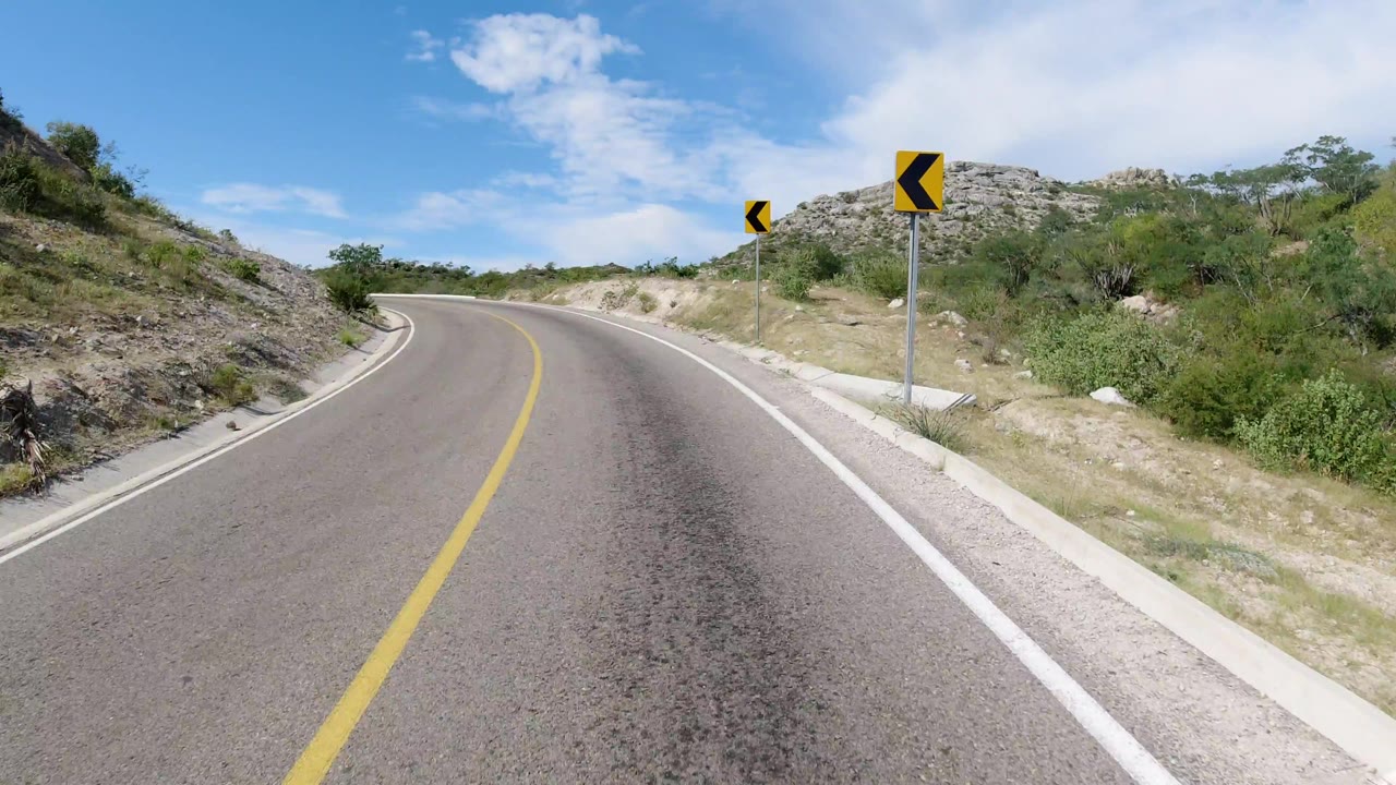 The road-level view in Los Cabos shows the curve drawing closer, with a black-and-yellow warning sign on the right shoulder and no visible people.