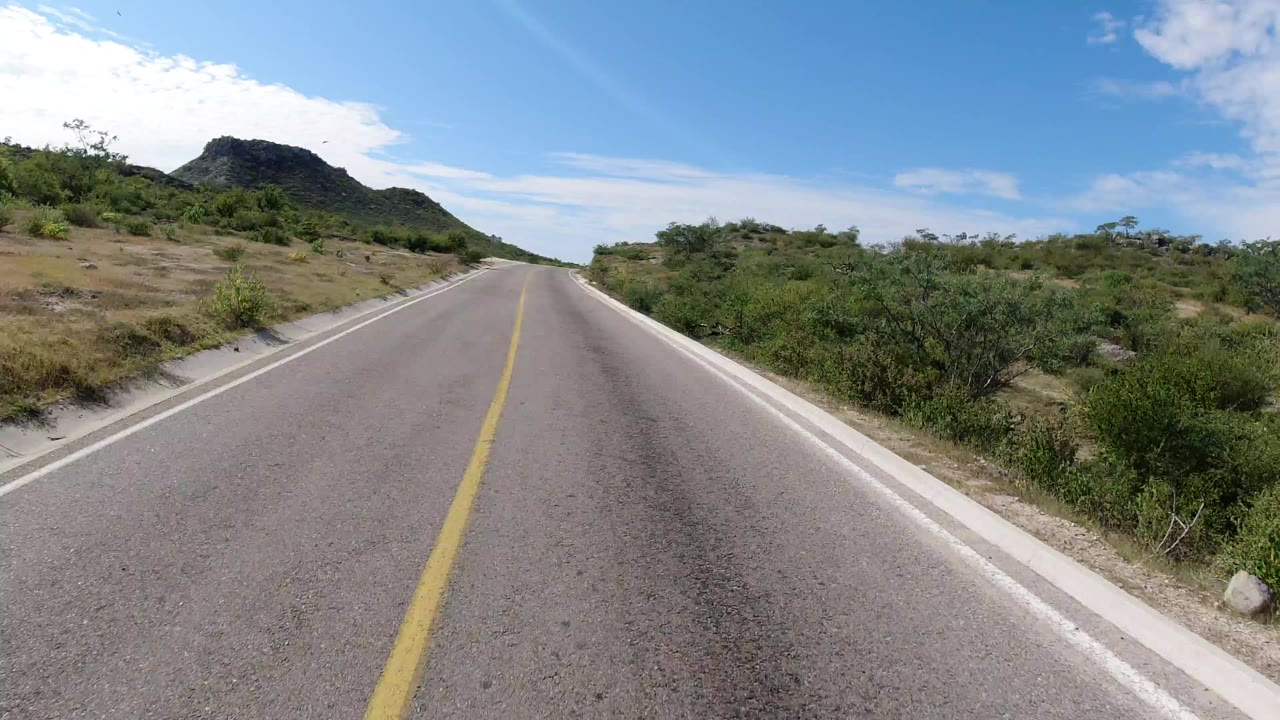 Empty paved road leading toward a low green hill under blue sky.