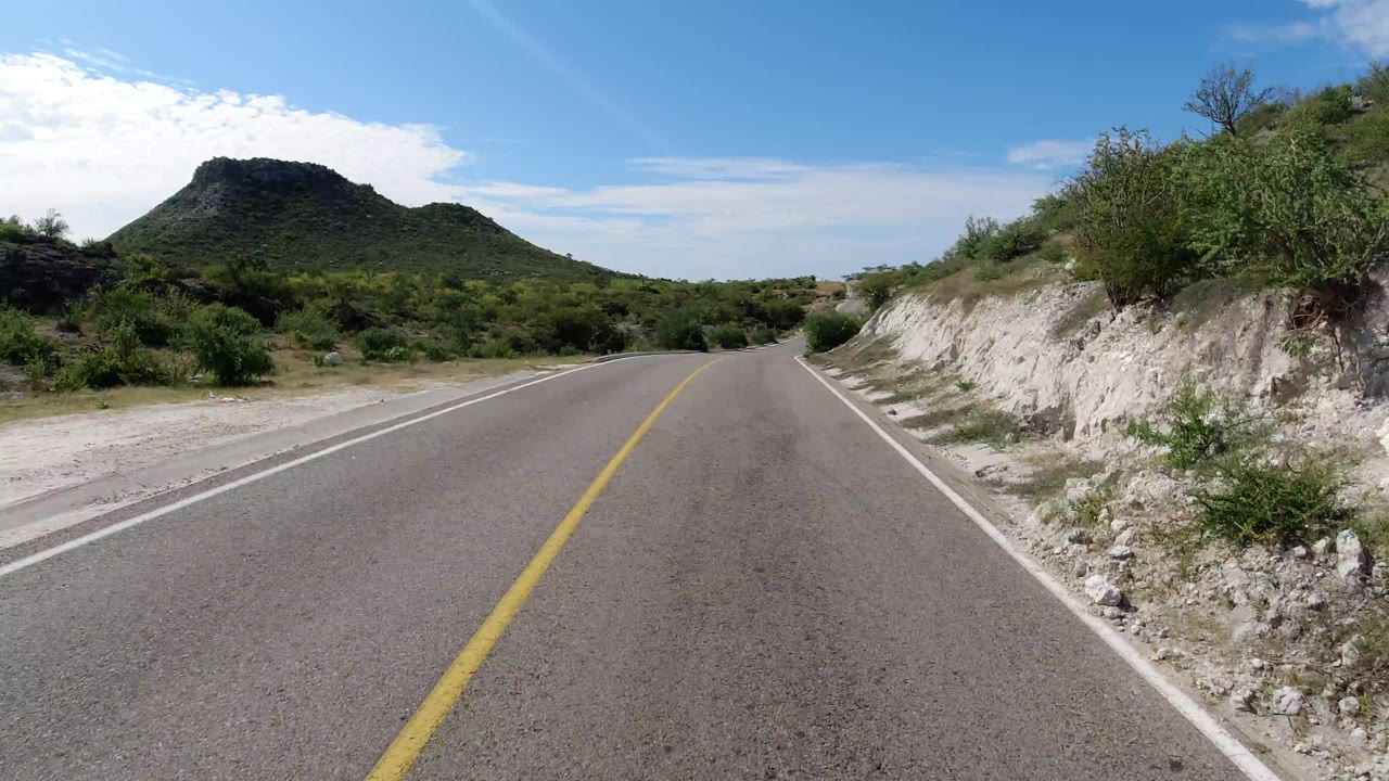 The road continues through Los Cabos, Mexico, bordered by pale rocky banks and dense green brush, with the dark rounded hill still centered in the distance and no visible people.