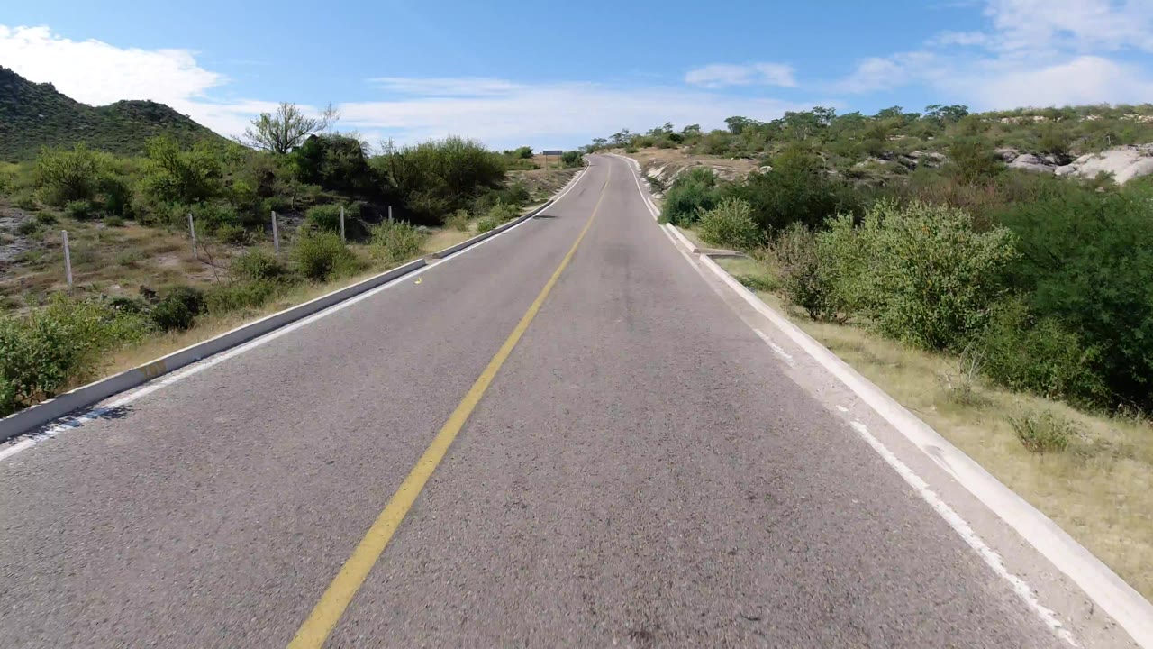 Start of the Los Cabos road clip, showing an empty two-lane paved road, yellow center line, dry brush on both sides, low hills, and no visible people.