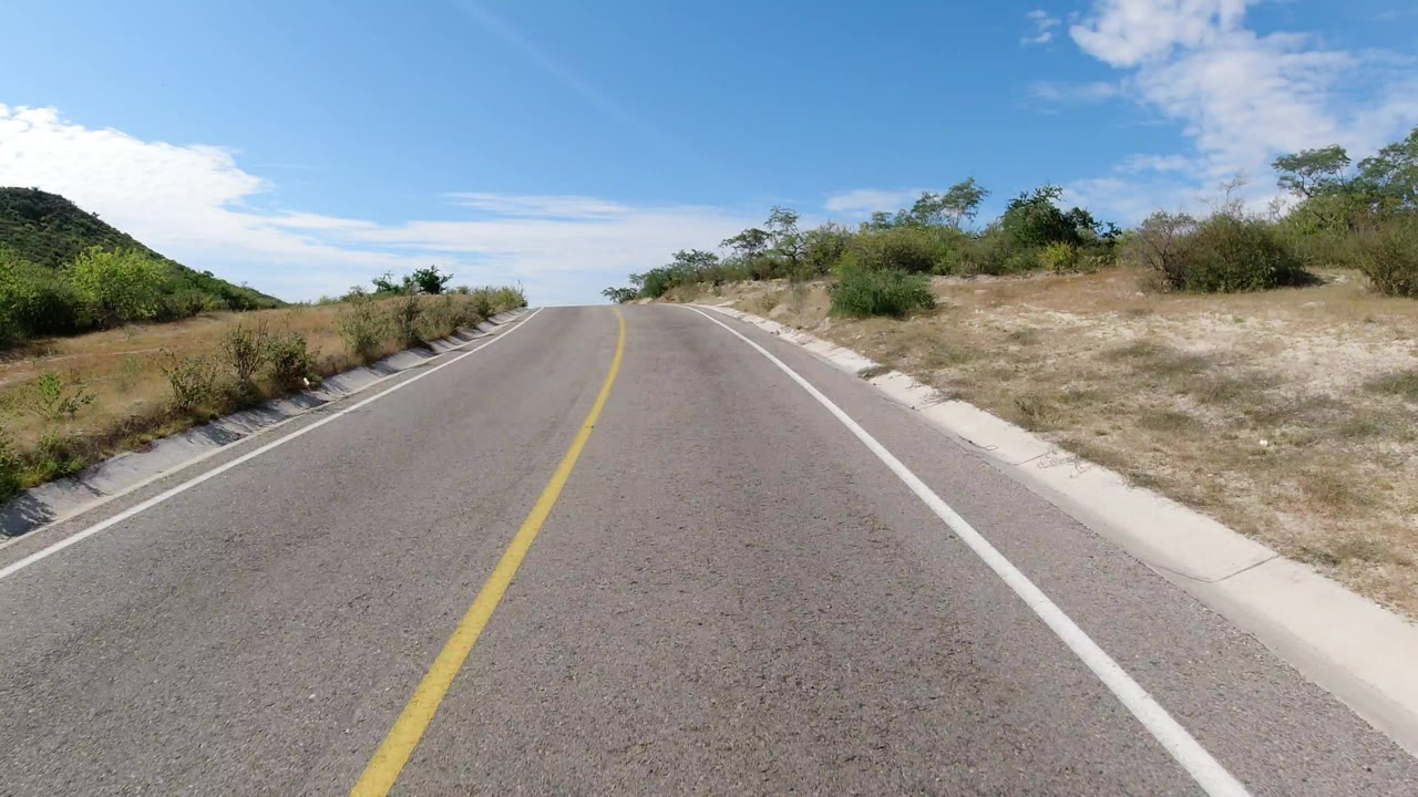 Two-lane road rising through dry roadside brush under blue sky.