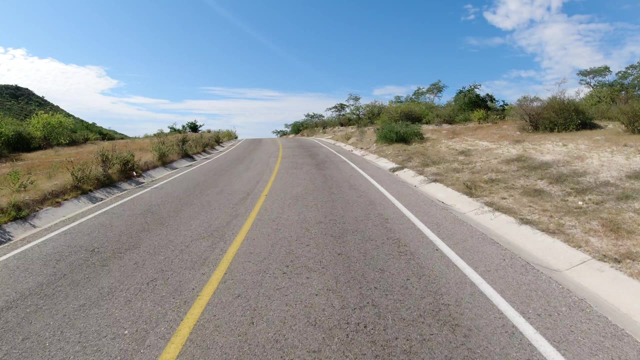The roadway continues through Los Cabos scrubland, with the yellow center line leading toward a small rise and no visible people.