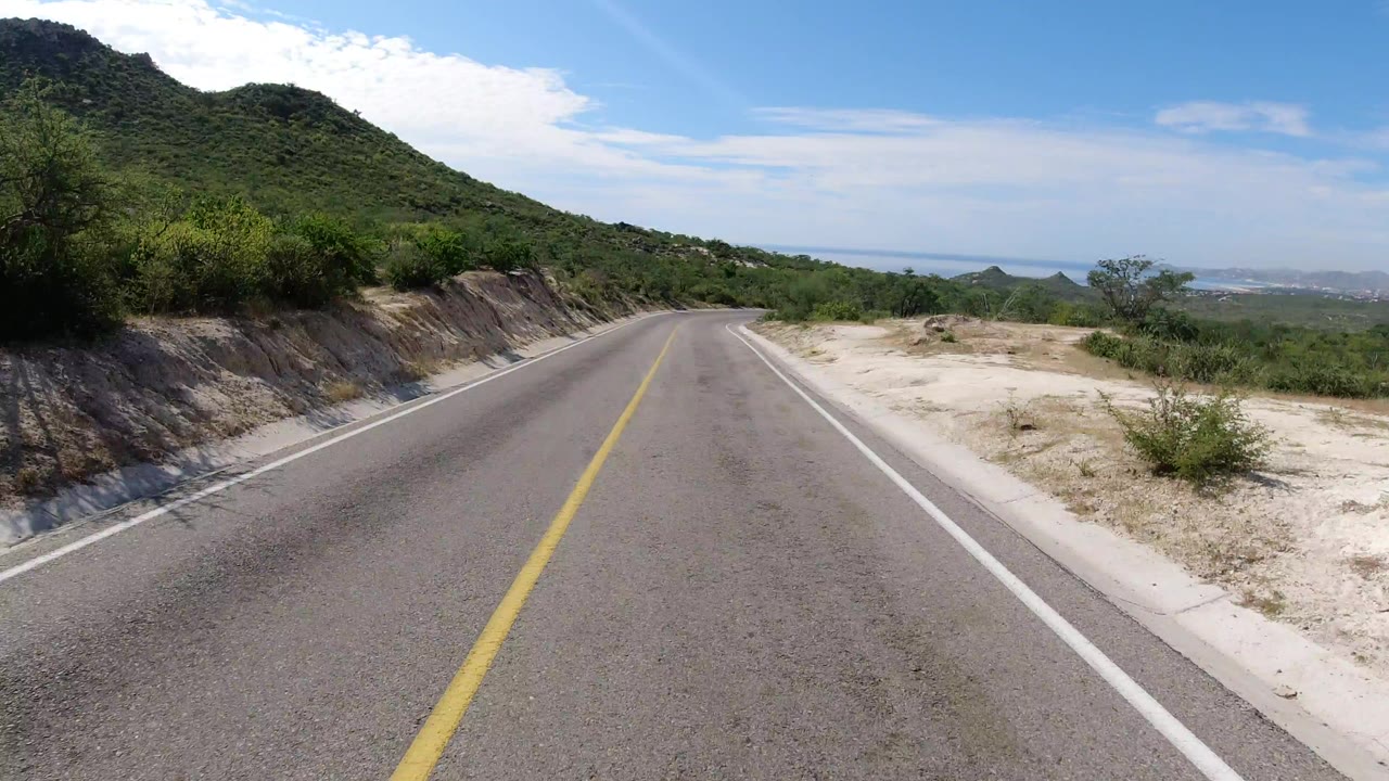 Empty two-lane road in Los Cabos with a yellow center line, sandy shoulder, green hillside, blue sky, distant water, and no visible people.