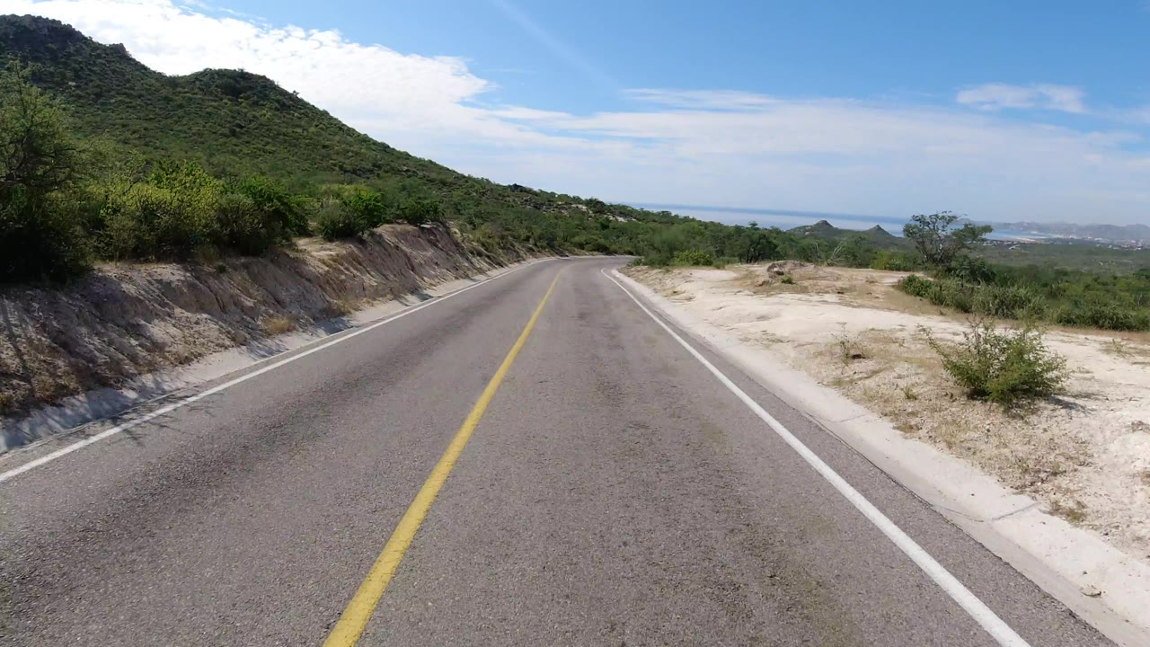 Forward view along a Los Cabos highway bordered by pale sand, roadside brush, a green hill on the left, distant coastline, and no visible people.