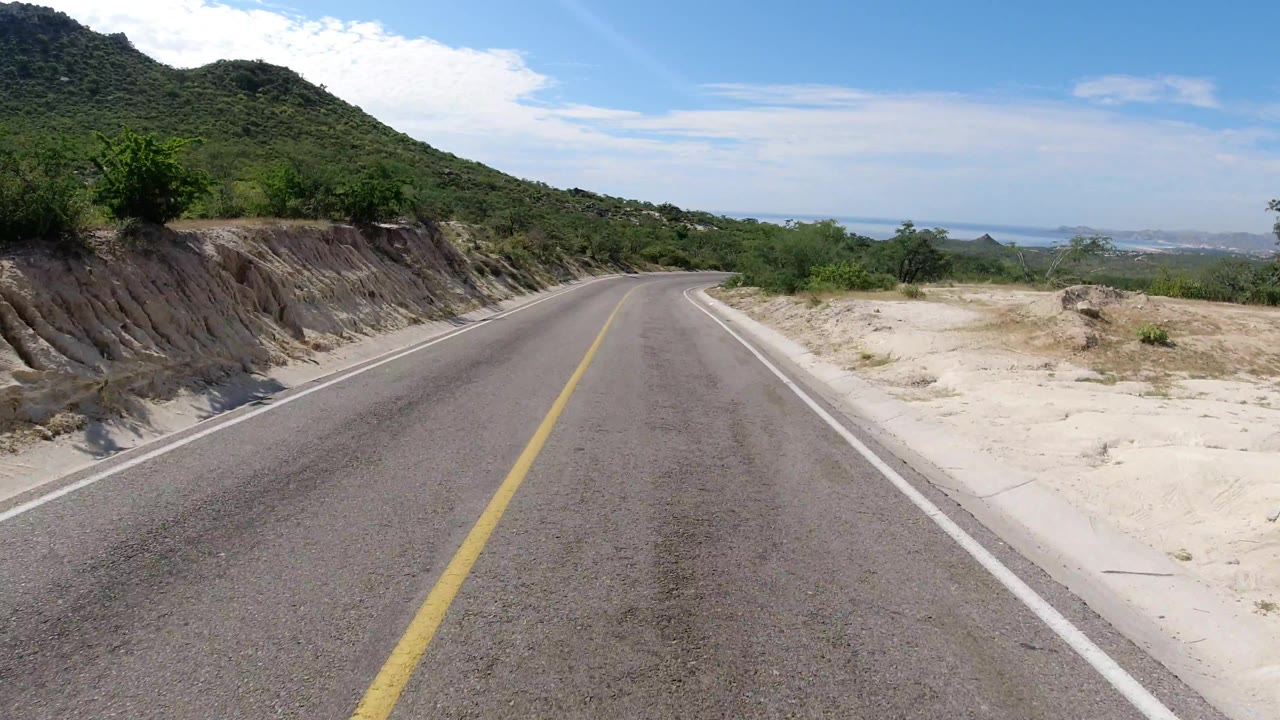 Curving highway bordered by hillside cut and pale sandy roadside.