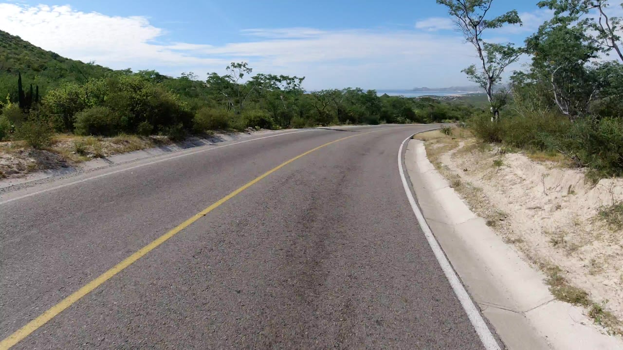 Start of the Los Cabos, Mexico clip showing a curving paved one-way road with sandy shoulder, green roadside brush, low hills, blue sky, and no visible people.