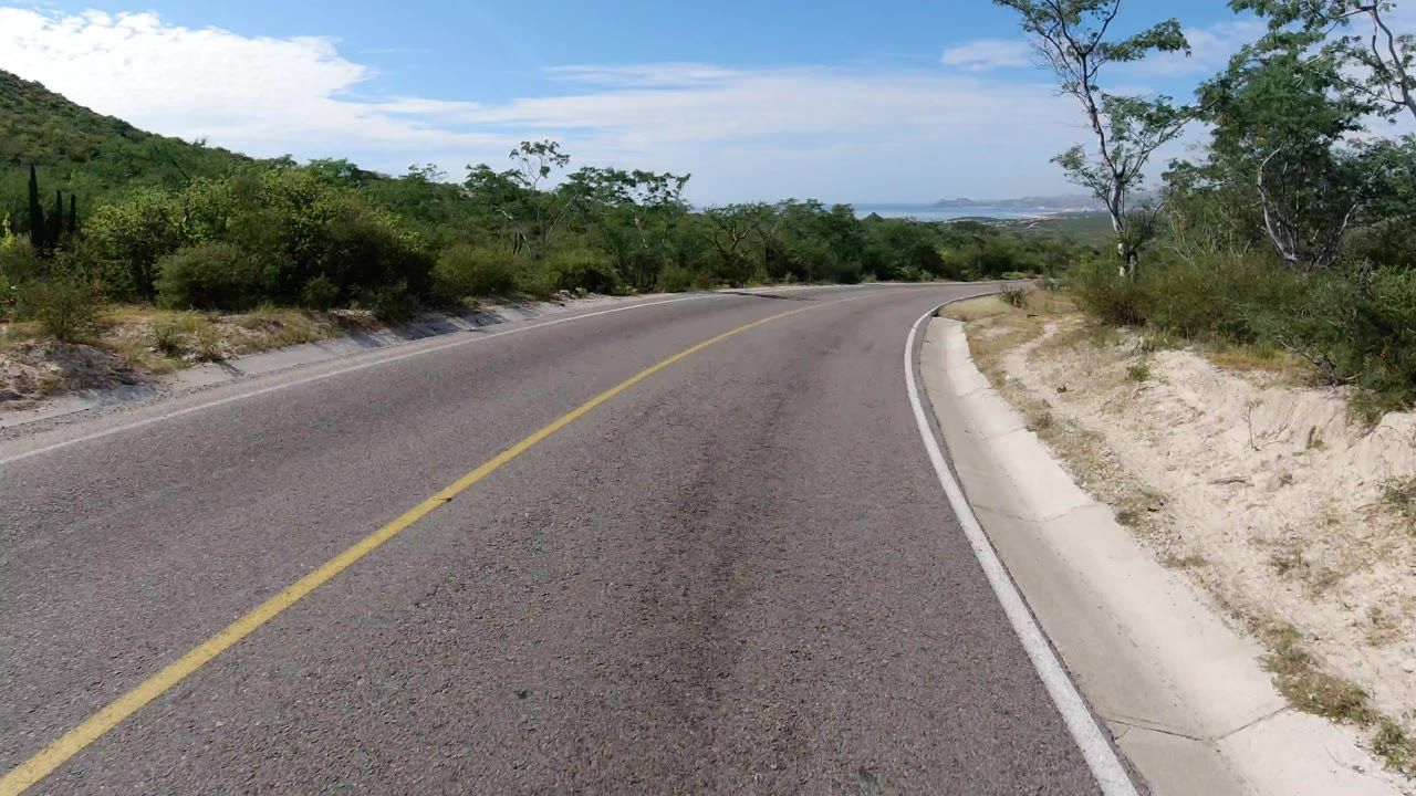 Los Cabos road view one second in, with the yellow centerline leading around a bend, dense green brush on both sides, pale sandy ground, and no visible people.