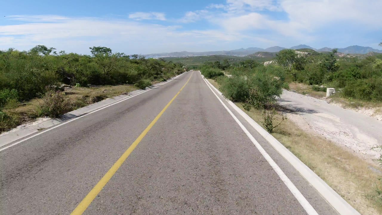 Start of the Los Cabos, Mexico road clip, showing a centered two-lane paved road, dry grass, green scrub, distant hills, bright sky, and no visible people.