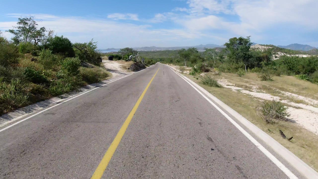 Forward road view in Los Cabos, Mexico with a yellow center line, sandy shoulders, green shrubs, distant hills, and no visible people.