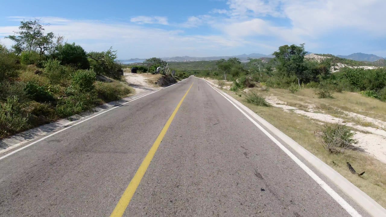 Centered road perspective in Los Cabos, Mexico with low roadside brush, a pale dirt shoulder, distant ridges, light cloud cover, and no visible people.