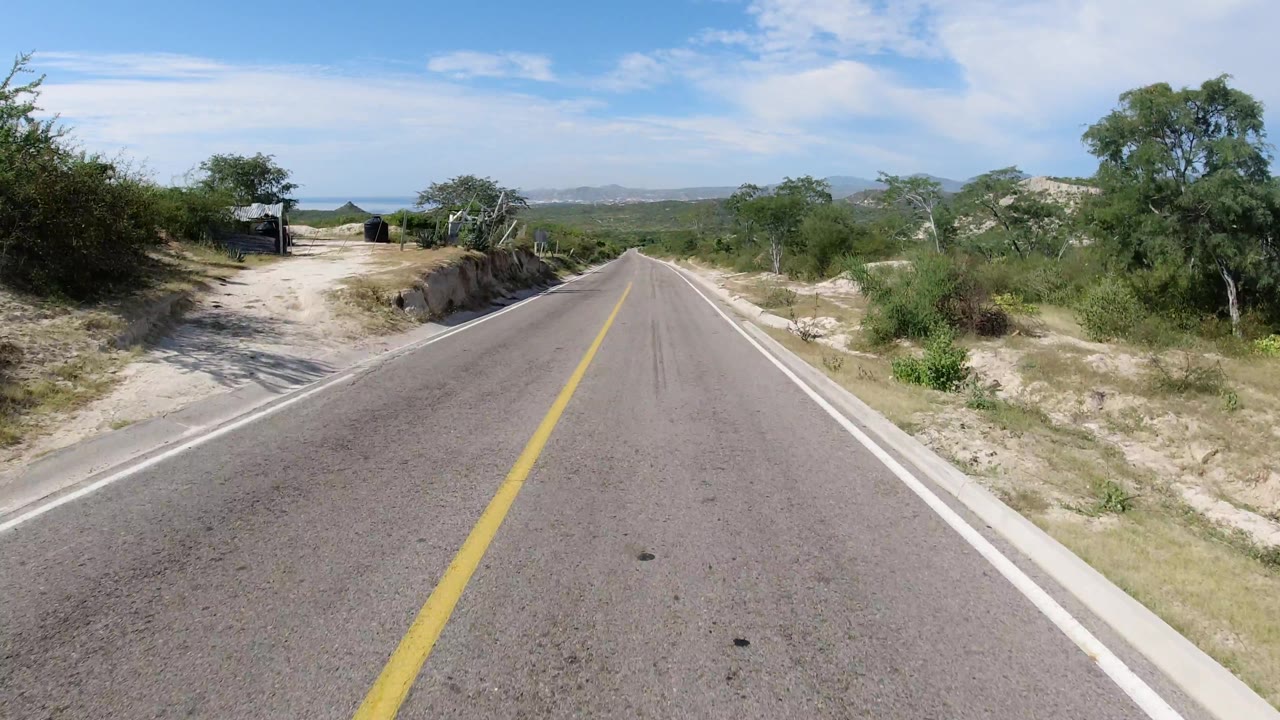 Road beside sandy embankments, bushes, and a small roadside structure.