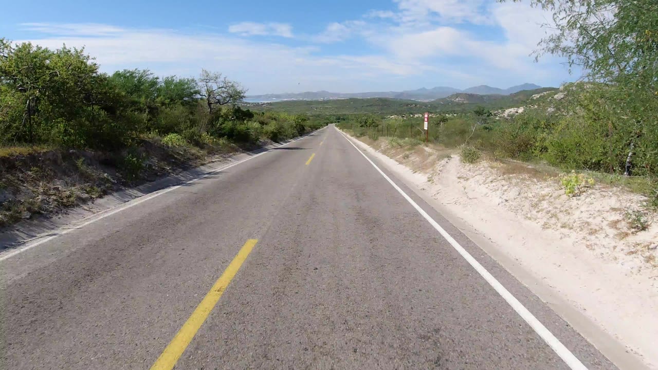 Forward-facing view down an empty paved road in Los Cabos, Mexico, with sandy shoulders, low green brush, distant hills, and no visible people.