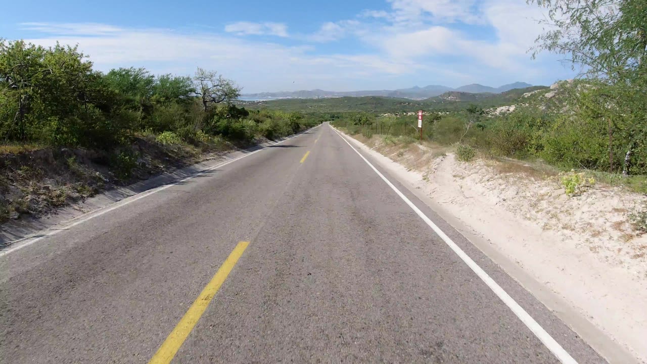 Centered roadway perspective in Los Cabos, Mexico, with a yellow center line, roadside brush, pale sand, distant hills, and no visible people.