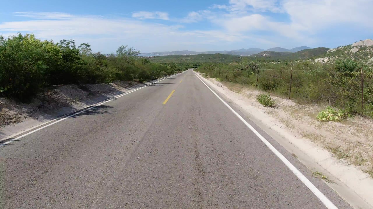 Start of a Los Cabos, Mexico road clip showing an empty two-lane highway descending between scrub, sandy shoulders, distant hills, and no visible people.