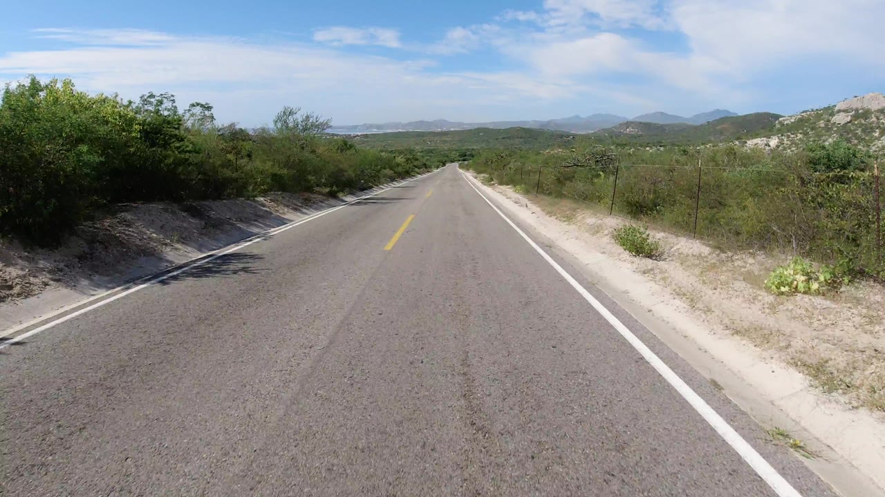 Los Cabos, Mexico highway view with the road centered, green roadside shrubs on both sides, low mountains on the horizon, and no visible people.