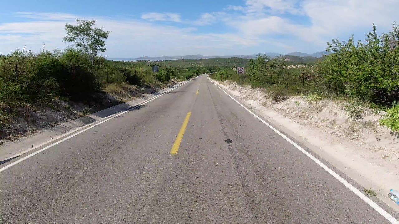 Opening view of an empty Los Cabos road with a yellow center line, sandy shoulders, green brush, distant hills, and no visible people.