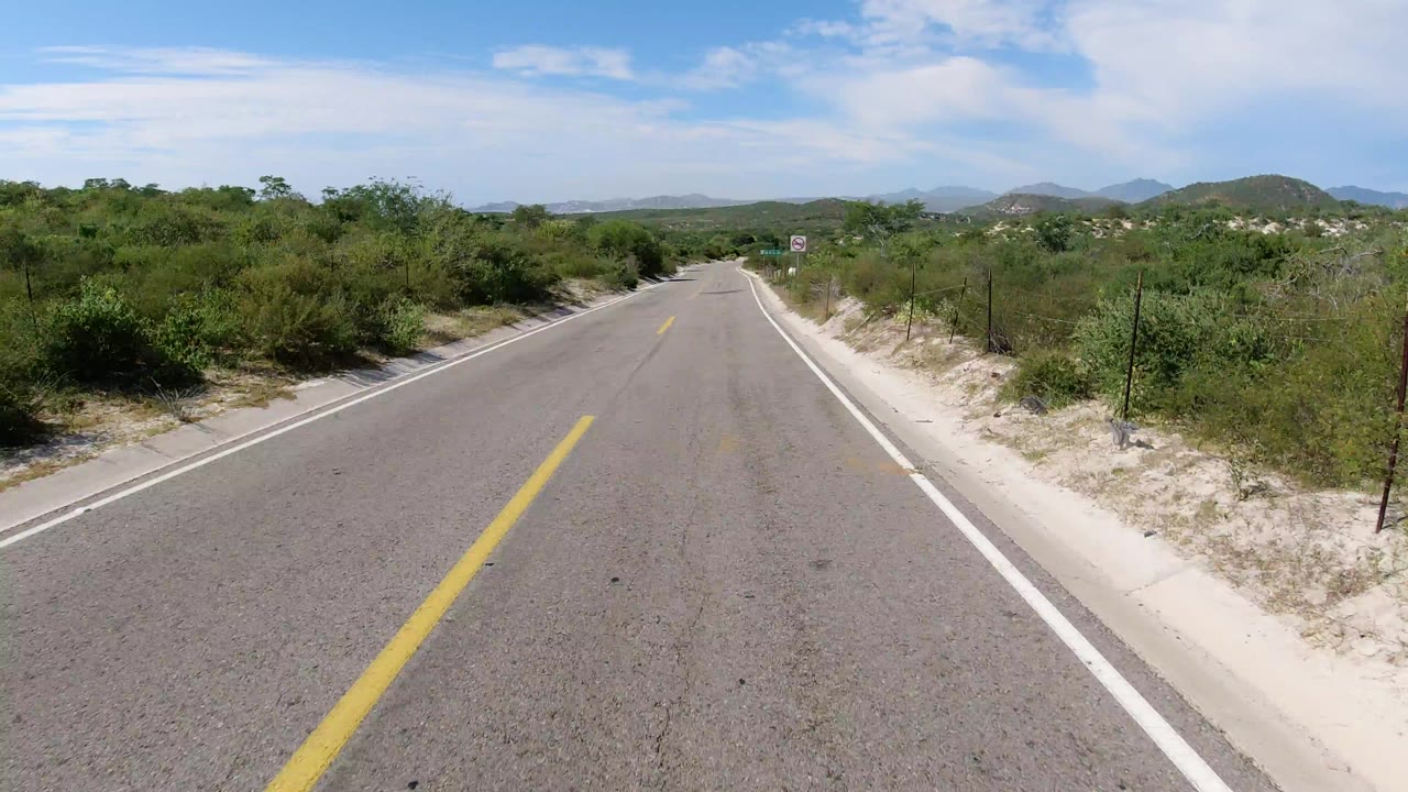 A centered view down an empty paved road in Los Cabos, Mexico, with scrub vegetation, sandy shoulders, distant hills, and no visible people.